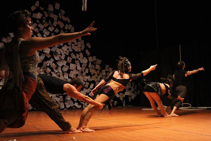 Three pairs of acrobats dancing on stage. In each pair, one acrobat leans impossibly forward extending one arm out, while their partner acrobat supports their legs so they can achieve this position. The lights are moody and the background looks as if hundreds of letters are stuck to the wall