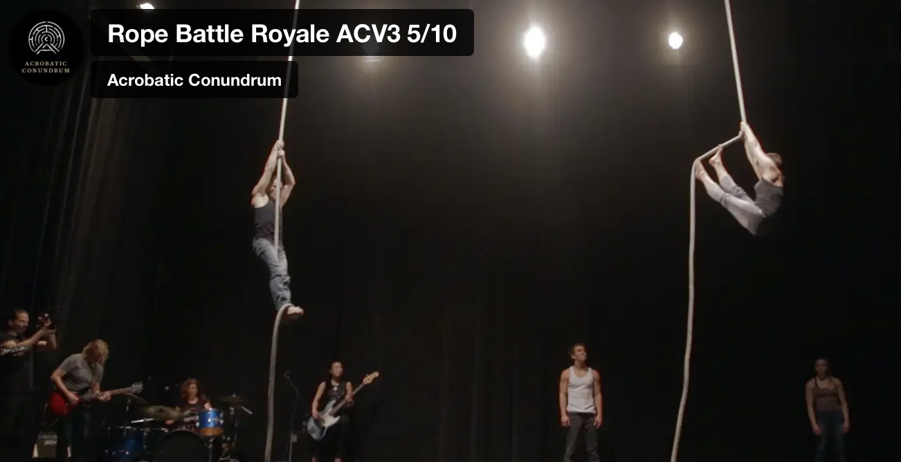 A bare black stage with stark lights. A rock band plays in the background to the left and two rope artists hang from ropes center stage while two other artists in the background look on.