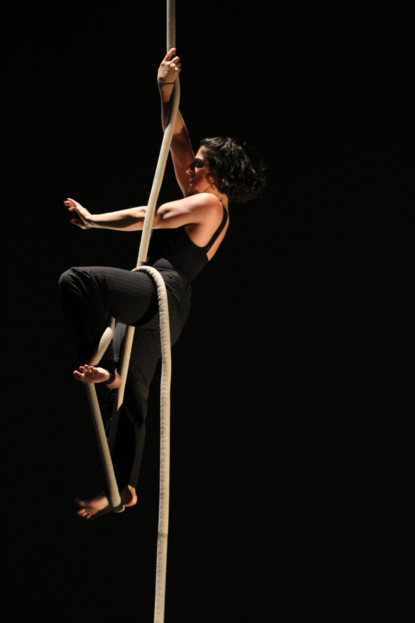 Me in an all black costume, climbing the rope. One arm reaches high while the other extends in front of me. Both feet are flexed as if climbing stairs.