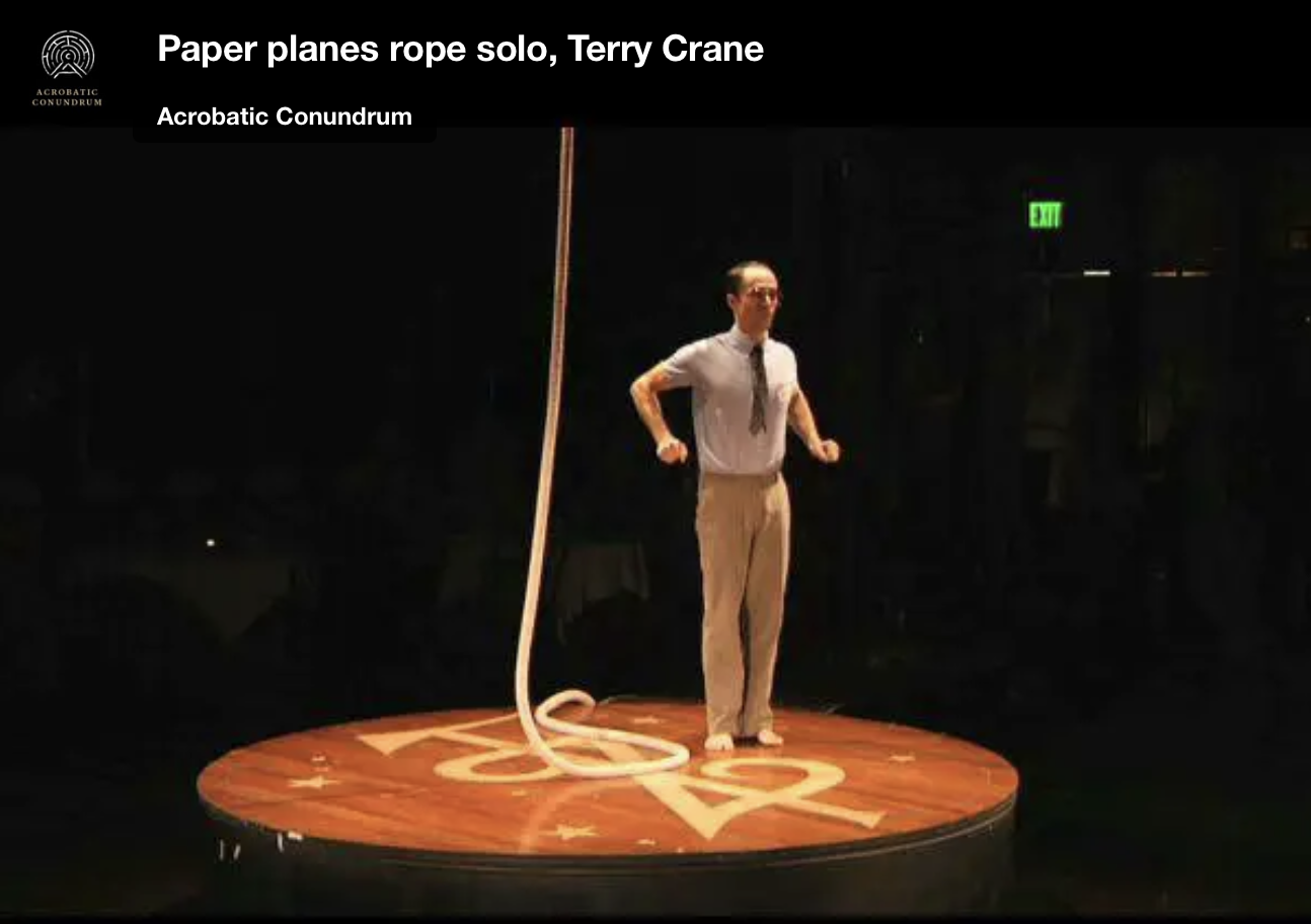 A man dressed as a stereotypical nerd in white, tucked-in button down, khaki pants, tight tie, and glasses. He&rsquo;s standing center stage next to a rope with his shoulders and elbows slightly raised as if he&rsquo;s very nervous.