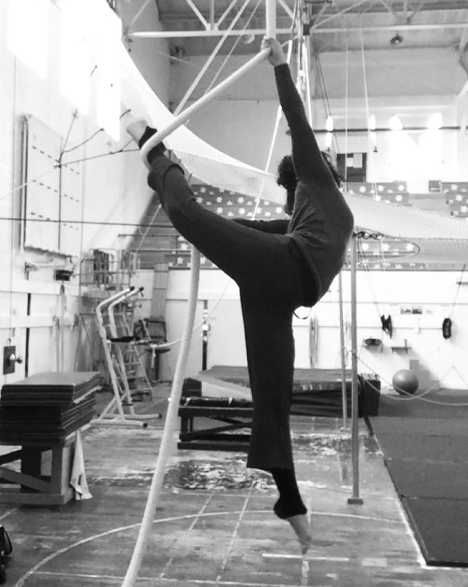 Black and white. I hang from a rope in a circus gymnasium. One arm hold the rope above my head while the other olds it out to the side. One of my leg is pulled up with the rope behind, between my hands, while the other extends to the floor. My body arches gently creating a dancer-like position. In the background are many mats and other apparatuses over a wooden floor. 