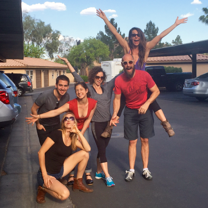 A group of six fit young 20-somethings stand in their hotel parking lot smiling at the camera in the hot Vegas sun. Many have their arms extended, all have big wide smiles or funny faces. One woman has climbed onto one of the male acrobat&rsquo;s backs and smiles from there.